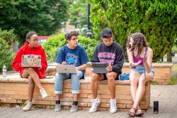 Four app sitting on a low brick wall on a sunny green quad. One girl is holding up her phone and gesturing toward it while the other three, using their laptops, look over at what she's showing them.